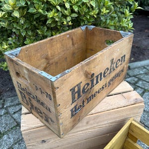 May include: A weathered wooden crate, branded with the Heineken logo in black lettering. The crate features metal corner protectors and hand-cut handles. The wood has a natural, aged look. The crate is placed on top of another wooden crate.