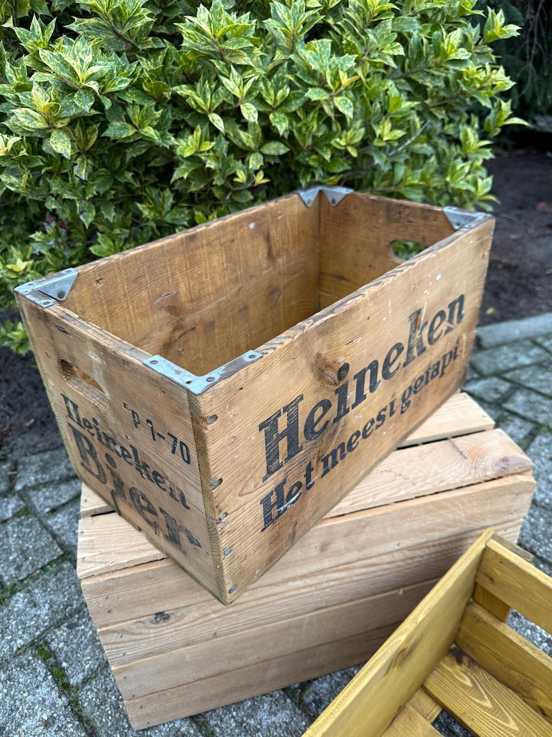 May include: A weathered wooden crate, branded with the Heineken logo in black lettering. The crate features metal corner protectors and hand-cut handles. The wood has a natural, aged look. The crate is placed on top of another wooden crate.