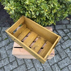 May include: A rectangular wooden crate with a light brown finish, featuring a slatted base and open sides. The crate is placed on top of a smaller, unfinished wooden pallet. Green and yellow foliage is in the background.