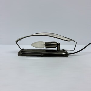Może przedstawiać: Czarna metalowa lampa biurkowa z zakrzywionym ramieniem i białą żar&oacute;wką. Lampa ma czarny przew&oacute;d i czarną podstawę.