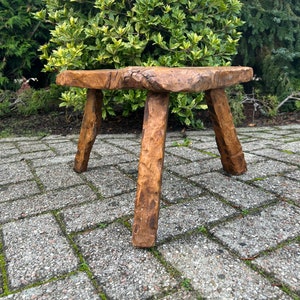 Puede incluir: Un taburete de madera r&uacute;stico con una tapa cuadrada y tres patas. El taburete est&aacute; hecho de madera toscamente labrada y tiene un acabado marr&oacute;n natural.