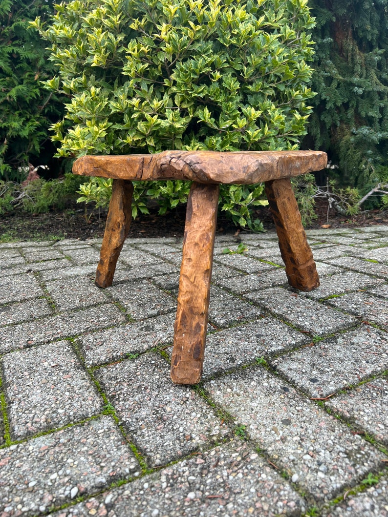 Puede incluir: Un taburete de madera r&uacute;stico con una tapa cuadrada y tres patas. El taburete est&aacute; hecho de madera toscamente labrada y tiene un acabado marr&oacute;n natural.