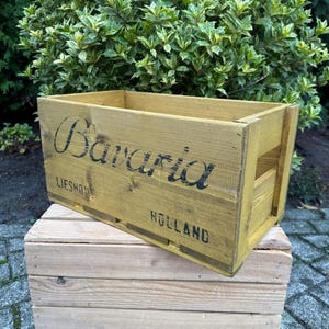 May include: A yellow-painted wooden crate with the word "Bavaria" in black lettering. It rests on a smaller, unpainted wooden crate. The text "Lieshout Holland" is also visible on the crate.