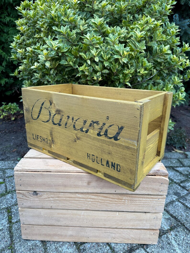 May include: A yellow-painted wooden crate with the word "Bavaria" in black lettering. It rests on a smaller, unpainted wooden crate. The text "Lieshout Holland" is also visible on the crate.
