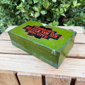 May include: A vintage green rectangular biscuit tin with the text "VERKADE'S NU ELCK SYN SIN BISCUITS" in black and red lettering. The tin is placed on a wooden crate, with green foliage in the background.