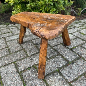 Puede incluir: Un taburete de madera r&uacute;stico con un acabado marr&oacute;n natural y tres patas. El taburete est&aacute; hecho de una sola pieza de madera y tiene una superficie rugosa y desigual.