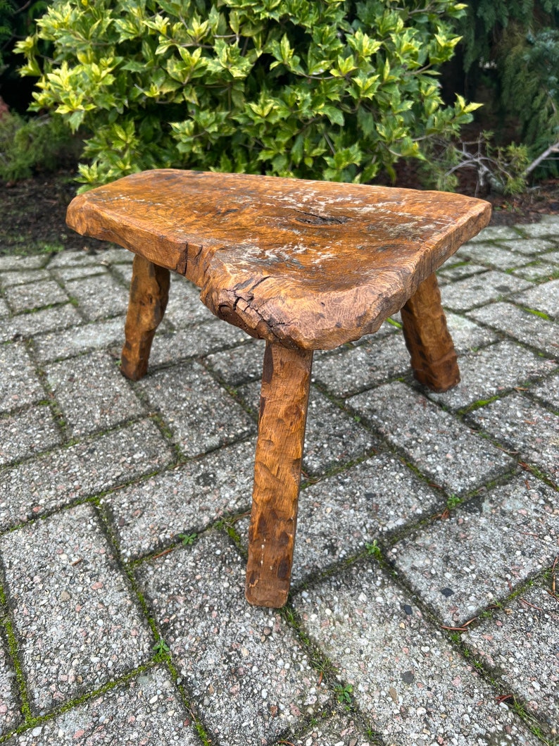 Puede incluir: Un taburete de madera r&uacute;stico con un acabado marr&oacute;n natural y tres patas. El taburete est&aacute; hecho de una sola pieza de madera y tiene una superficie rugosa y desigual.