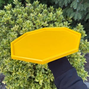 May include: A bright yellow, geometric tray with a multi-sided shape. The tray is held against a backdrop of green foliage. The tray's edges are slightly raised, and the color is solid.