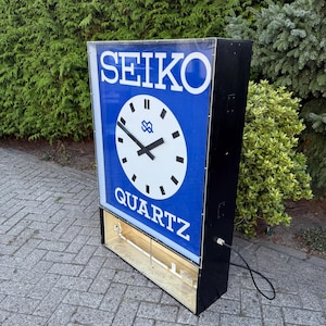 Könnte beinhalten: Vintage Seiko Quartz Uhrenschild mit blauem Zifferblatt und weißer Schrift. Das Schild zeigt ein Zifferblatt mit schwarzen Zeigern und dem Seiko-Logo. Das Schild ist in einem schwarzen rechteckigen Rahmen eingefasst.