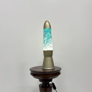 Könnte beinhalten: Eine tealfarbene und weiße Lavalampe mit goldener Basis steht auf einem dunkelbraunen Holzsockel.