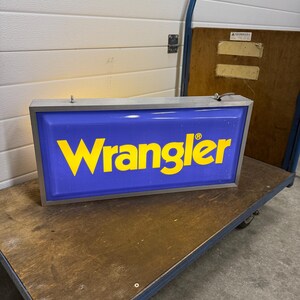 May include: A rectangular blue and yellow Wrangler sign with a silver frame. The word "Wrangler" is in yellow, bold letters. The sign is on a wooden cart with blue metal framing.