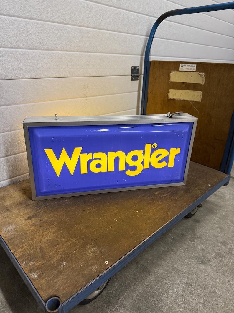 May include: A rectangular blue and yellow Wrangler sign with a silver frame. The word "Wrangler" is in yellow, bold letters. The sign is on a wooden cart with blue metal framing.