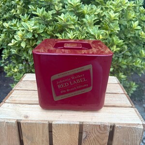 May include: A red, square ice bucket with a handle on top. The front of the bucket features a label with the text "Johnnie Walker RED LABEL Old Scotch Whisky." The bucket is sitting on a wooden crate.