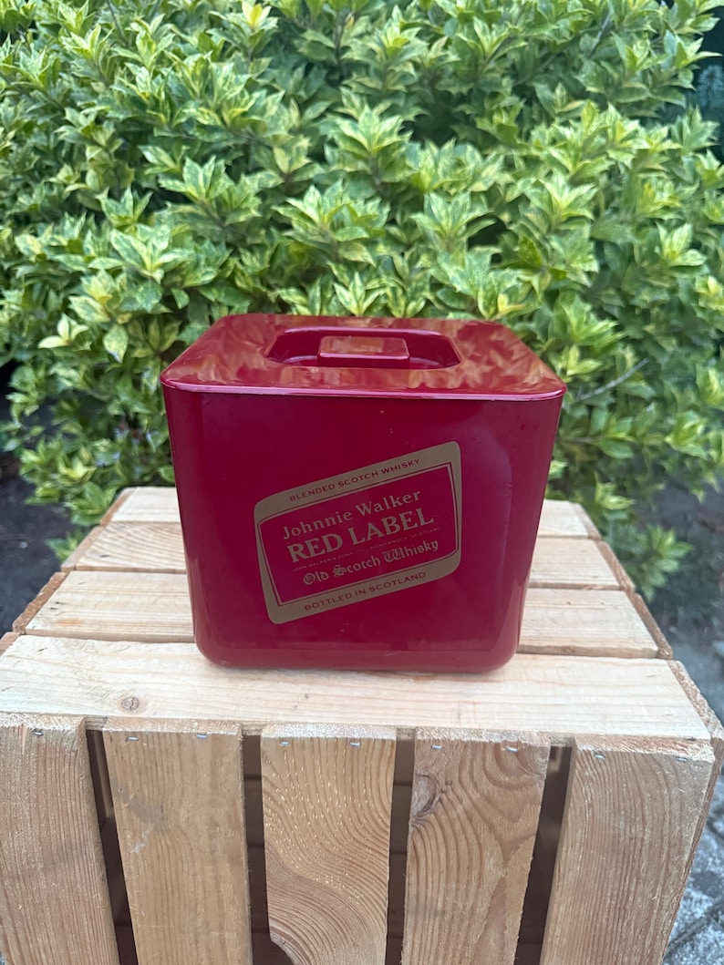 May include: A red, square ice bucket with a handle on top. The front of the bucket features a label with the text "Johnnie Walker RED LABEL Old Scotch Whisky." The bucket is sitting on a wooden crate.