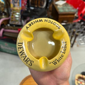 May include: Yellow ceramic ashtray with the text "DEWAR'S" and "Scotch Whisky White Label" in black. The ashtray has a circular design with three cigarette rests.