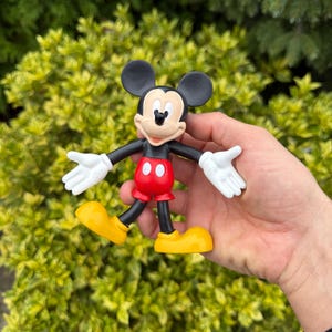 Pode incluir: Uma estatueta do Mickey Mouse. A figura tem orelhas pretas, um corpo preto, cal&ccedil;&otilde;es vermelhos com bot&otilde;es brancos, sapatos amarelos e luvas brancas. A figura &eacute; segurada em frente a um fundo de folhas verdes.