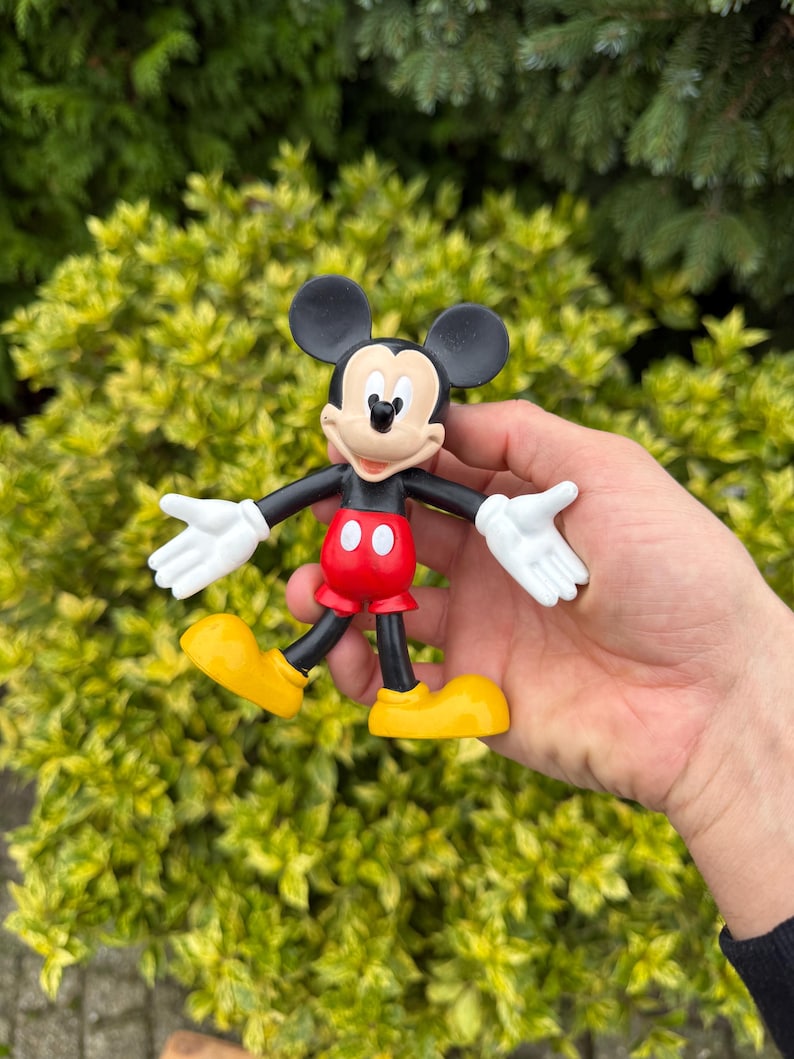 Pode incluir: Uma estatueta do Mickey Mouse. A figura tem orelhas pretas, um corpo preto, cal&ccedil;&otilde;es vermelhos com bot&otilde;es brancos, sapatos amarelos e luvas brancas. A figura &eacute; segurada em frente a um fundo de folhas verdes.