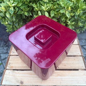 May include: A square, burgundy-colored plastic container with a recessed lid. The container sits on a wooden crate, with green foliage in the background. The container has a glossy finish.