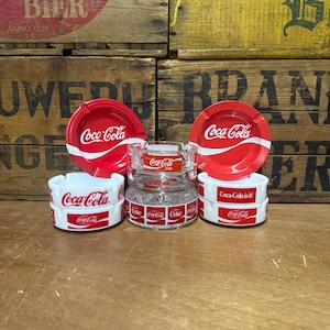 May include: A collection of five Coca-Cola branded ashtrays. Two are red with white Coca-Cola logos, two are white with red Coca-Cola logos and text, and one is a clear glass ashtray featuring multiple small Coca-Cola and Coke Light logos.