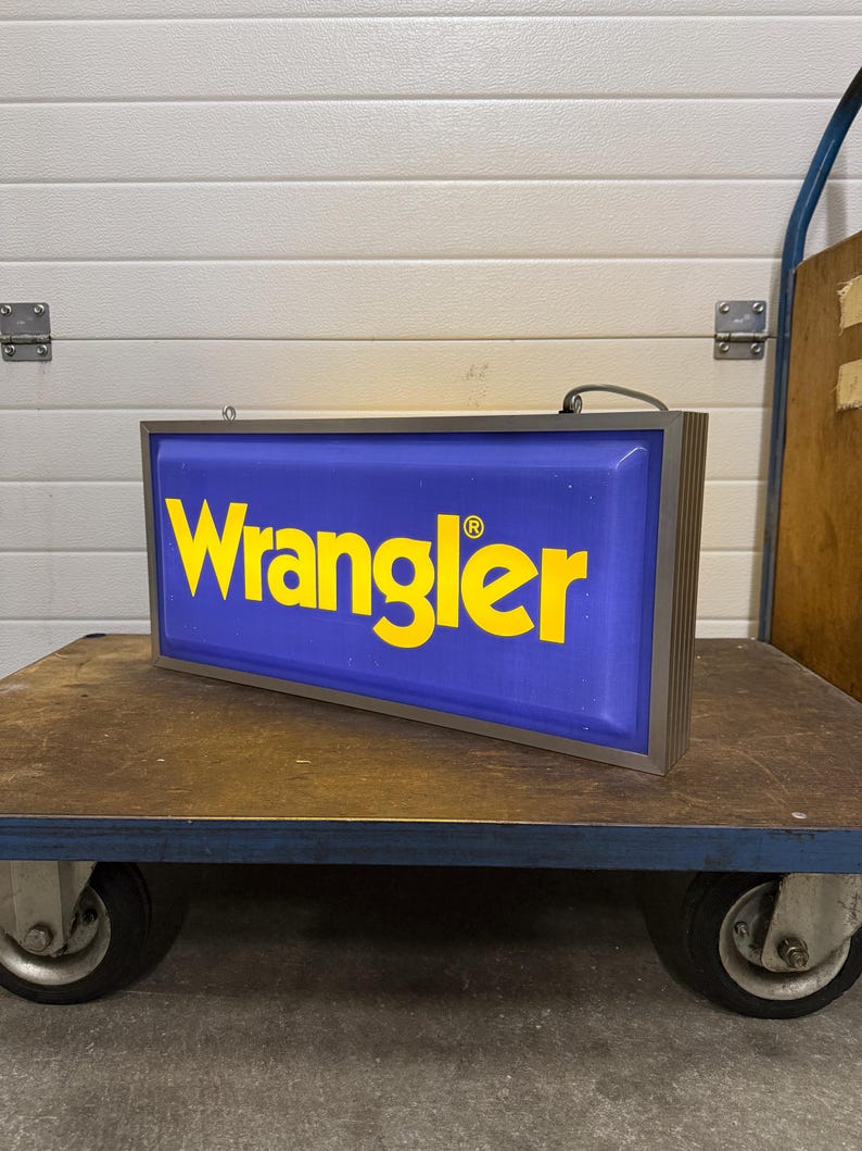May include: Rectangular blue and yellow Wrangler sign with a silver frame. The word "Wrangler" is in large yellow letters on a blue background. The sign is sitting on a brown cart with wheels.