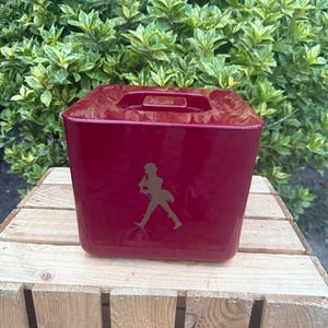 May include: A burgundy-colored, square ice bucket with a handle on top. The bucket features a gold-colored silhouette of a man walking. The ice bucket is sitting on a wooden crate, with green foliage in the background.