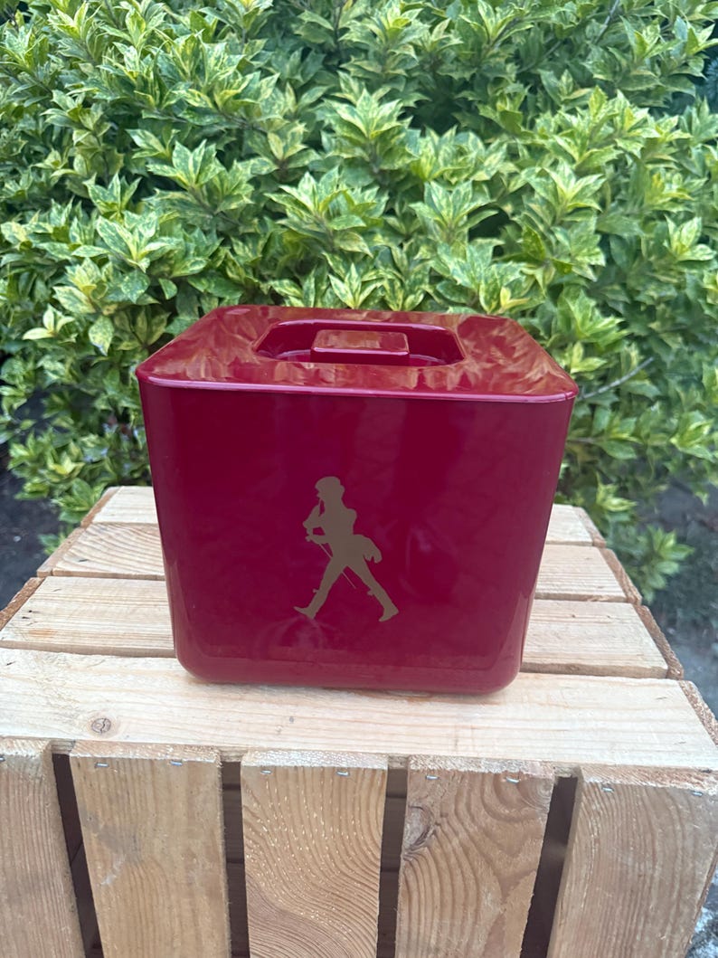 May include: A burgundy-colored, square ice bucket with a handle on top. The bucket features a gold-colored silhouette of a man walking. The ice bucket is sitting on a wooden crate, with green foliage in the background.