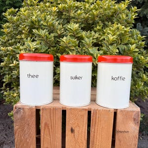 May include: Three white canisters with red lids, labeled "thee", "suiker", and "koffie" in black text. The canisters are arranged on a wooden crate, with green foliage in the background.