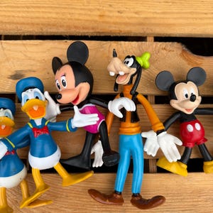 Pode incluir: Uma cole&ccedil;&atilde;o de figuras de personagens da Disney, incluindo Mickey Mouse, Minnie Mouse, Pato Donald e Pateta. As figuras s&atilde;o coloridas e posam em v&aacute;rias poses. As figuras s&atilde;o feitas de pl&aacute;stico e s&atilde;o exibidas em uma superf&iacute;cie de madeira.