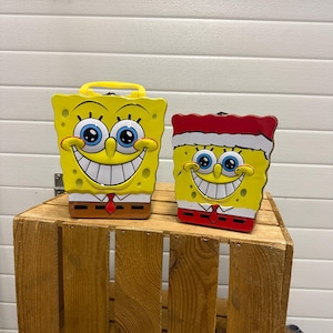 Pode incluir: Duas lancheiras amarelas do Bob Esponja com rostos sorridentes, olhos azuis e gravatas vermelhas. Uma tem uma alça amarela, a outra uma borda vermelha e branca. Elas estão em uma caixa de madeira.