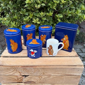 May include: A collection of blue metal storage tins and a white mug featuring cartoon bear illustrations. The tins vary in size and shape, with lids and a rectangular container. The mug has a handle. The items are arranged on a wooden crate.