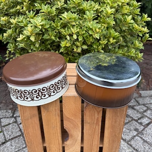 Op de afbeelding: Twee vintage metalen cakeblikken. Eén is bruin en wit met een decoratief patroon, de andere is bruin met een zilveren rand. Ze staan op een houten krat, met groene bladeren op de achtergrond.