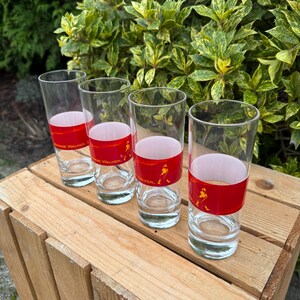 May include: Four clear glass tumblers with a red band featuring the text "Johnnie Walker" and a silhouette of a man. The glasses are arranged on a wooden crate, with green foliage in the background.