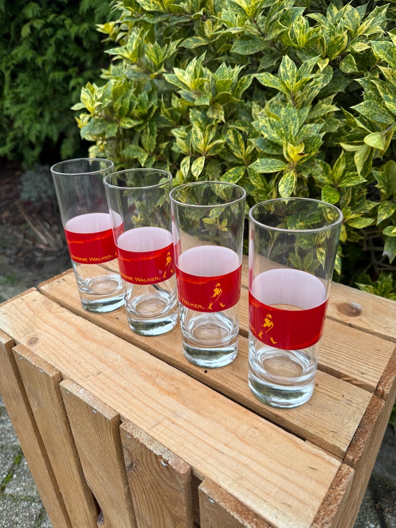 May include: Four clear glass tumblers with a red band featuring the text "Johnnie Walker" and a silhouette of a man. The glasses are arranged on a wooden crate, with green foliage in the background.