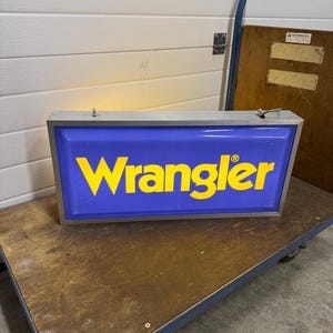 May include: A rectangular, illuminated sign with the word "Wrangler" in yellow letters on a blue background. The sign has a silver frame and is resting on a wooden cart with wheels. The sign is likely for advertising.