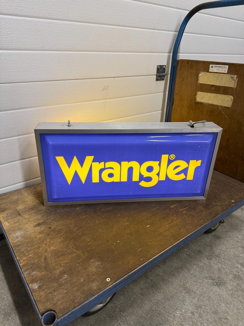 May include: A rectangular, illuminated sign with the word "Wrangler" in yellow letters on a blue background. The sign has a silver frame and is resting on a wooden cart with wheels. The sign is likely for advertising.