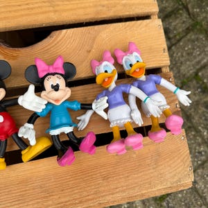 Pode incluir: Uma cole&ccedil;&atilde;o de figuras de personagens da Disney, incluindo Minnie Mouse e Margarida, dispostas numa caixa de madeira. Minnie Mouse usa um vestido azul e um la&ccedil;o rosa. Margarida usa um top roxo e um la&ccedil;o rosa. As figuras s&atilde;o coloridas.