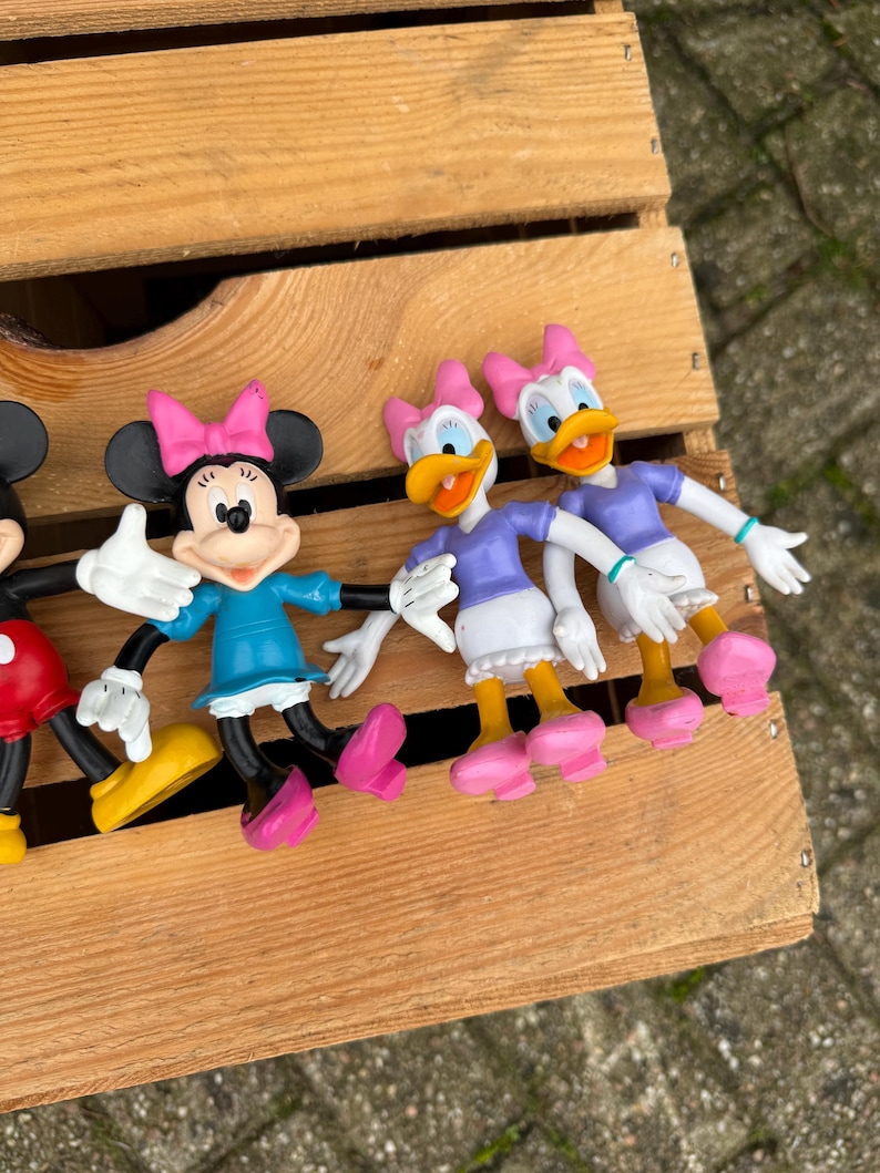 Pode incluir: Uma cole&ccedil;&atilde;o de figuras de personagens da Disney, incluindo Minnie Mouse e Margarida, dispostas numa caixa de madeira. Minnie Mouse usa um vestido azul e um la&ccedil;o rosa. Margarida usa um top roxo e um la&ccedil;o rosa. As figuras s&atilde;o coloridas.