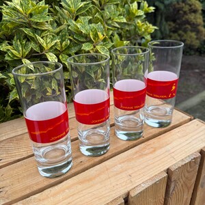 May include: Four clear highball glasses with a red band featuring the text "Johnnie Walker" and a logo. The glasses are arranged on a wooden surface, with green foliage in the background.