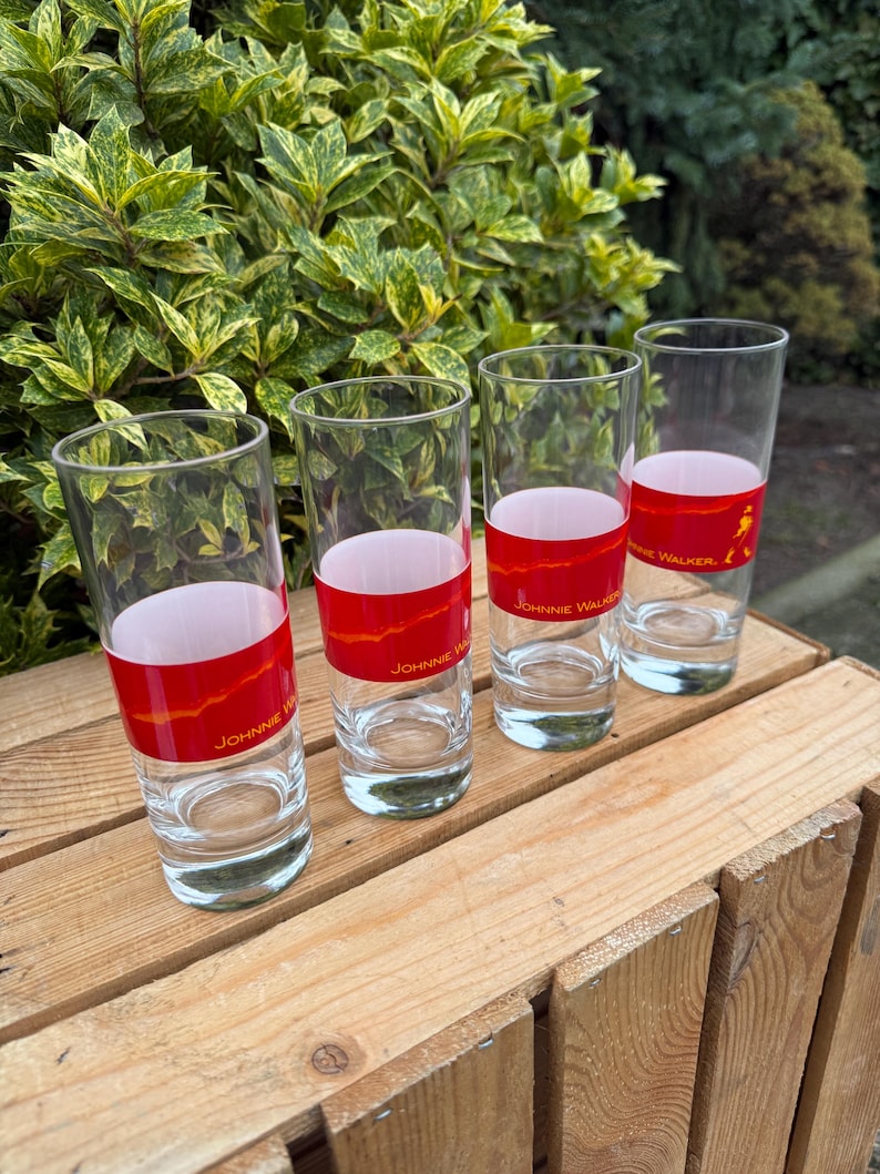 May include: Four clear highball glasses with a red band featuring the text "Johnnie Walker" and a logo. The glasses are arranged on a wooden surface, with green foliage in the background.