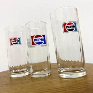 Puede incluir: Tres vasos de beber de vidrio transparente con un diseño retorcido. Cada vaso tiene una pegatina de logotipo de Pepsi roja y azul.
