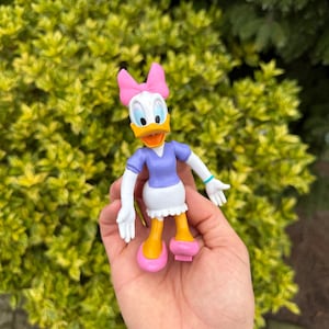 Pode incluir: Uma figura da Margarida, com rosto branco, bico laranja e la&ccedil;o rosa. Ela usa um top roxo, saia branca e sapatos rosa. A figura &eacute; segurada em uma m&atilde;o contra um fundo de folhagem verde.