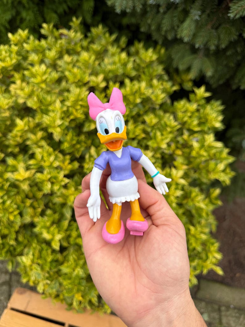 Pode incluir: Uma figura da Margarida, com rosto branco, bico laranja e la&ccedil;o rosa. Ela usa um top roxo, saia branca e sapatos rosa. A figura &eacute; segurada em uma m&atilde;o contra um fundo de folhagem verde.