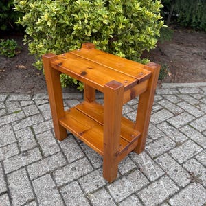 Op de afbeelding: Kleine, houten bijzettafel met twee niveaus en een warme, honingkleurige afwerking. De tafel heeft een rechthoekig blad en een onderste plank, ondersteund door vier stevige poten. De houtnerf is zichtbaar.
