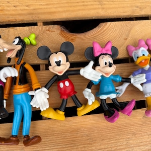 Pode incluir: Uma cole&ccedil;&atilde;o de figuras de personagens da Disney, incluindo Mickey Mouse, Minnie Mouse, Pateta e Margarida. Mickey usa cal&ccedil;as vermelhas e sapatos amarelos. Minnie usa um vestido azul e um la&ccedil;o rosa. Pateta tem um colete laranja e cal&ccedil;as azuis. Margarida usa um top roxo e um la&ccedil;o rosa.
