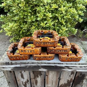 May include: A stack of six brown ceramic ashtrays with yellow and orange accents. The ashtrays are decorated with the words "Hotel du Centre".