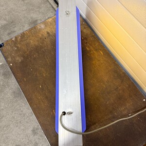 May include: A rectangular, vertical sign with a blue center and silver metal sides. A power cord extends from the bottom. The sign is resting on a wooden surface.