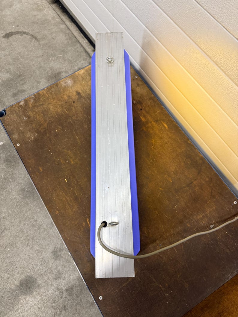 May include: A rectangular, vertical sign with a blue center and silver metal sides. A power cord extends from the bottom. The sign is resting on a wooden surface.