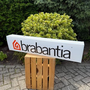 May include: A white rectangular sign with the word "brabantia" in black text and a red and orange logo. The sign is resting on a wooden crate, with green foliage in the background.