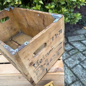May include: A vintage wooden crate with metal corner brackets. The crate features cut-out handles and the text "Heineken Beer" printed on the side. It is a rectangular shape and is placed on top of another wooden crate.