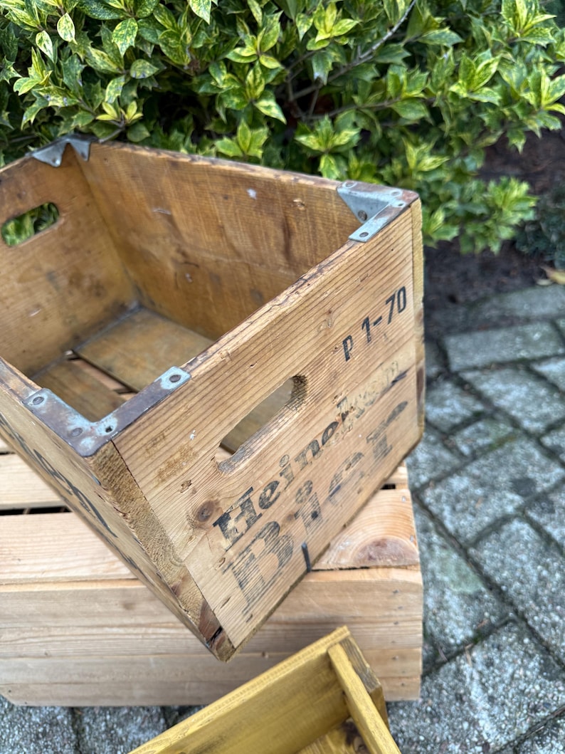 May include: A vintage wooden crate with metal corner brackets. The crate features cut-out handles and the text "Heineken Beer" printed on the side. It is a rectangular shape and is placed on top of another wooden crate.